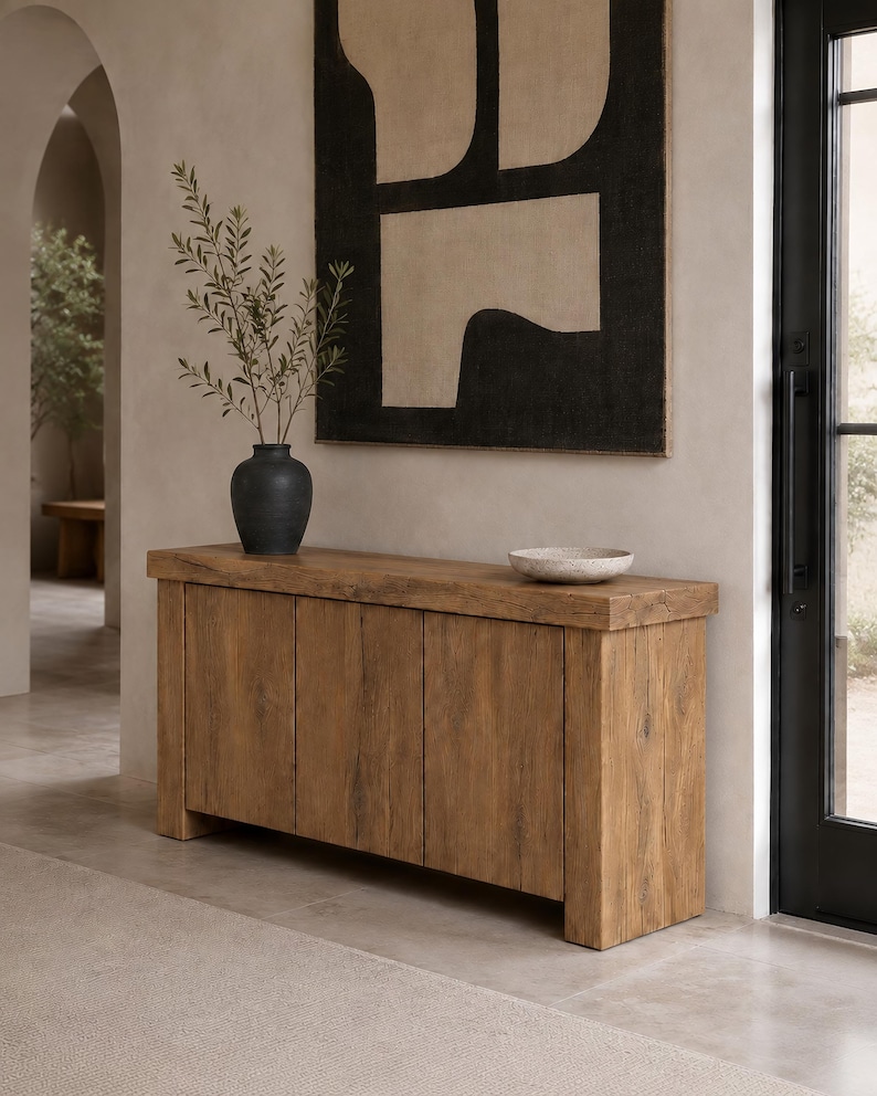 Peut inclure: Une console rectangulaire en bois avec un plateau &eacute;pais et quatre portes verticales pr&eacute;sentant une finition au grain naturel. Un vase en c&eacute;ramique gris anthracite contenant des branches feuillues et un petit bol en pierre mouchet&eacute;e.