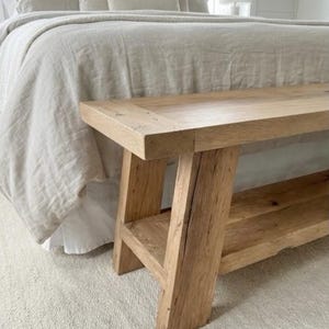 May include: A natural wood bench with a rectangular top and a lower shelf. The bench has four sturdy legs and is placed in front of a bed with neutral-colored bedding. The wood has visible grain and knots.