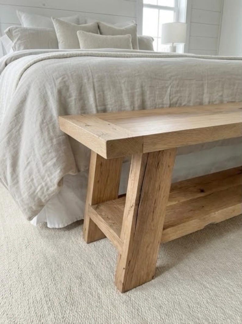 May include: A natural wood bench with a rectangular top and a lower shelf. The bench has four sturdy legs and is placed in front of a bed with neutral-colored bedding. The wood has visible grain and knots.