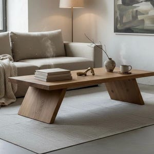 Puede incluir: Sal&oacute;n moderno con mesa de centro de madera clara con patas anguladas. La mesa tiene libros, un objeto decorativo, un jarr&oacute;n con una ramita y una taza. Un sof&aacute; beige, una alfombra y obras de arte completan la decoraci&oacute;n minimalista.