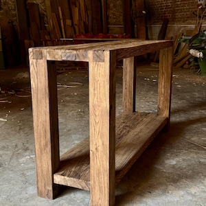 Peut inclure: Table console rectangulaire en bois rustique avec une &eacute;tag&egrave;re inf&eacute;rieure. Fabriqu&eacute;e &agrave; partir de planches &eacute;paisses et patin&eacute;es, elle r&eacute;v&egrave;le le grain et les n&oelig;uds naturels du bois. Les pieds et le cadre carr&eacute;s lui conf&egrave;rent un aspect robuste et artisanal. Dimensions: environ 120 cm de long.