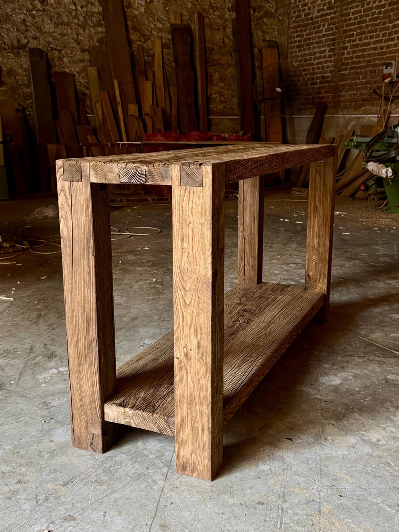 Peut inclure: Table console rectangulaire en bois rustique avec une &eacute;tag&egrave;re inf&eacute;rieure. Fabriqu&eacute;e &agrave; partir de planches &eacute;paisses et patin&eacute;es, elle r&eacute;v&egrave;le le grain et les n&oelig;uds naturels du bois. Les pieds et le cadre carr&eacute;s lui conf&egrave;rent un aspect robuste et artisanal. Dimensions: environ 120 cm de long.