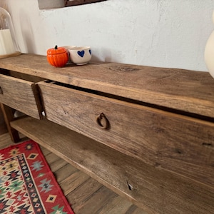 May include: A rustic wooden console table with two drawers and a lower shelf. The table has a natural wood finish and metal ring pulls. Decorative items on top include a small orange pumpkin, a white bowl with blue hearts, and a white ceramic vase.
