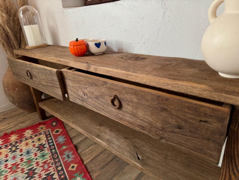 May include: A rustic wooden console table with two drawers and a lower shelf. The table has a natural wood finish and metal ring pulls. Decorative items on top include a small orange pumpkin, a white bowl with blue hearts, and a white ceramic vase.