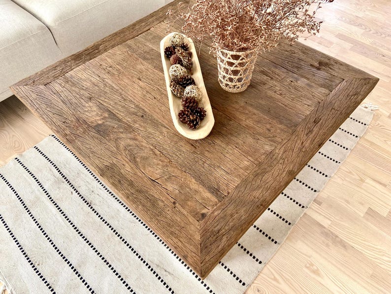 May include: A square, rustic wooden coffee table with a distressed finish. A long, cream-colored wooden bowl filled with pine cones sits on the table. A woven basket with dried flowers is also on the table. A striped rug is beneath the table.