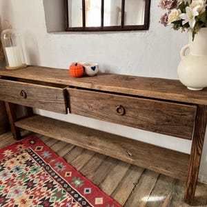 May include: A rustic wooden console table with two drawers and a lower shelf. The table has a natural wood finish and metal ring pulls. A vase of flowers, decorative pumpkins, and a rug are in the foreground.