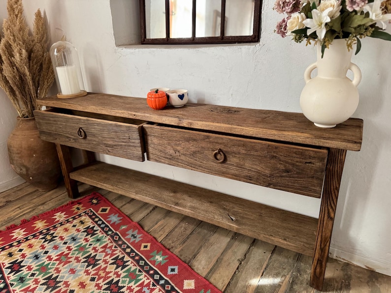 May include: A rustic wooden console table with two drawers and a lower shelf. The table has a natural wood finish and metal ring pulls. A vase of flowers, decorative pumpkins, and a rug are in the foreground.