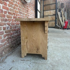 May include: A light-colored wooden cabinet with three drawers, featuring a natural wood grain finish. The cabinet has a rectangular shape with decorative brackets and a slightly overhanging top. The cabinet is set against a brick wall.