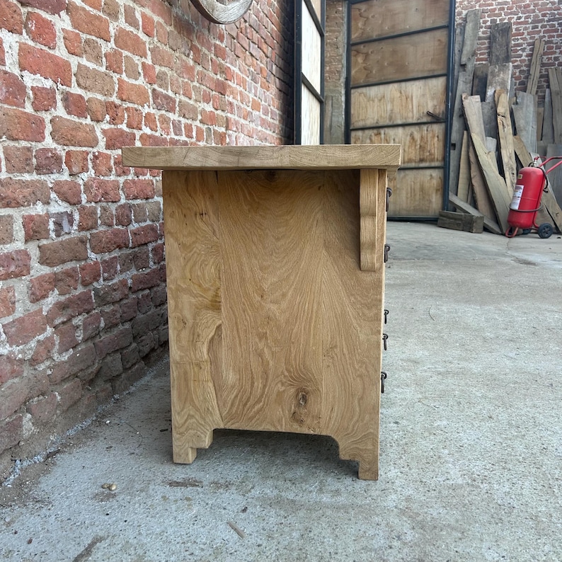 May include: A light-colored wooden cabinet with three drawers, featuring a natural wood grain finish. The cabinet has a rectangular shape with decorative brackets and a slightly overhanging top. The cabinet is set against a brick wall.