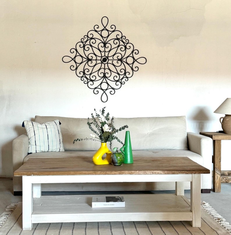 May include: A white sofa with a black metal wall decoration above it. A coffee table with a white base and a wooden top sits in front of the sofa. There are two vases on the table, one yellow and one green. A book is on the lower shelf of the table.