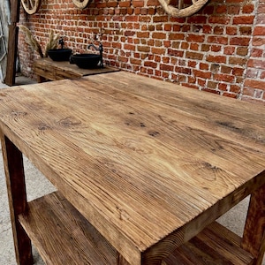 May include: A rustic wooden table with a single shelf. The table has a natural wood finish and is made of reclaimed wood. The table is in front of a brick wall with three wooden wagon wheels hanging on the wall.