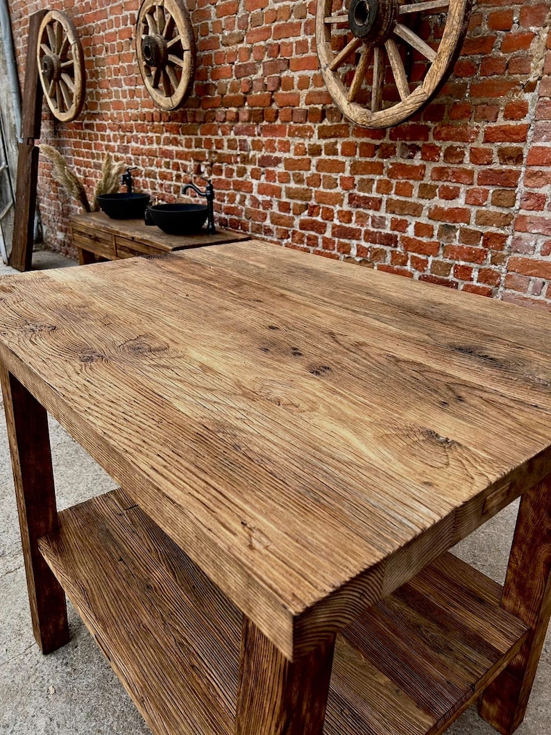 May include: A rustic wooden table with a single shelf. The table has a natural wood finish and is made of reclaimed wood. The table is in front of a brick wall with three wooden wagon wheels hanging on the wall.