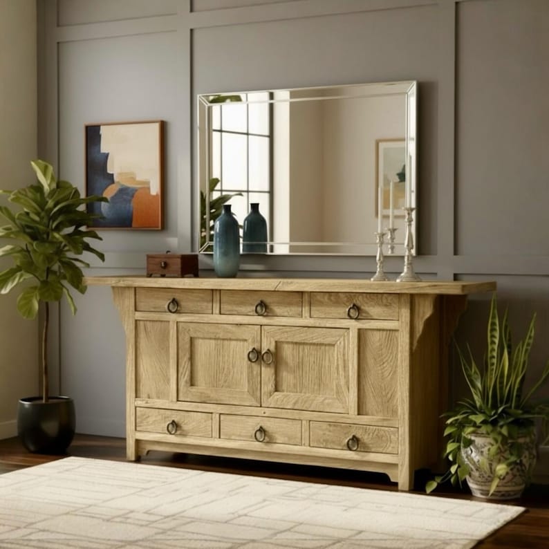 May include: A light-colored wooden sideboard with multiple drawers and cabinet doors, adorned with dark metal ring pulls. A large rectangular mirror hangs above the sideboard, reflecting the room. Decorative vases and a painting are on the sideboard. Potted plants flank the furniture.