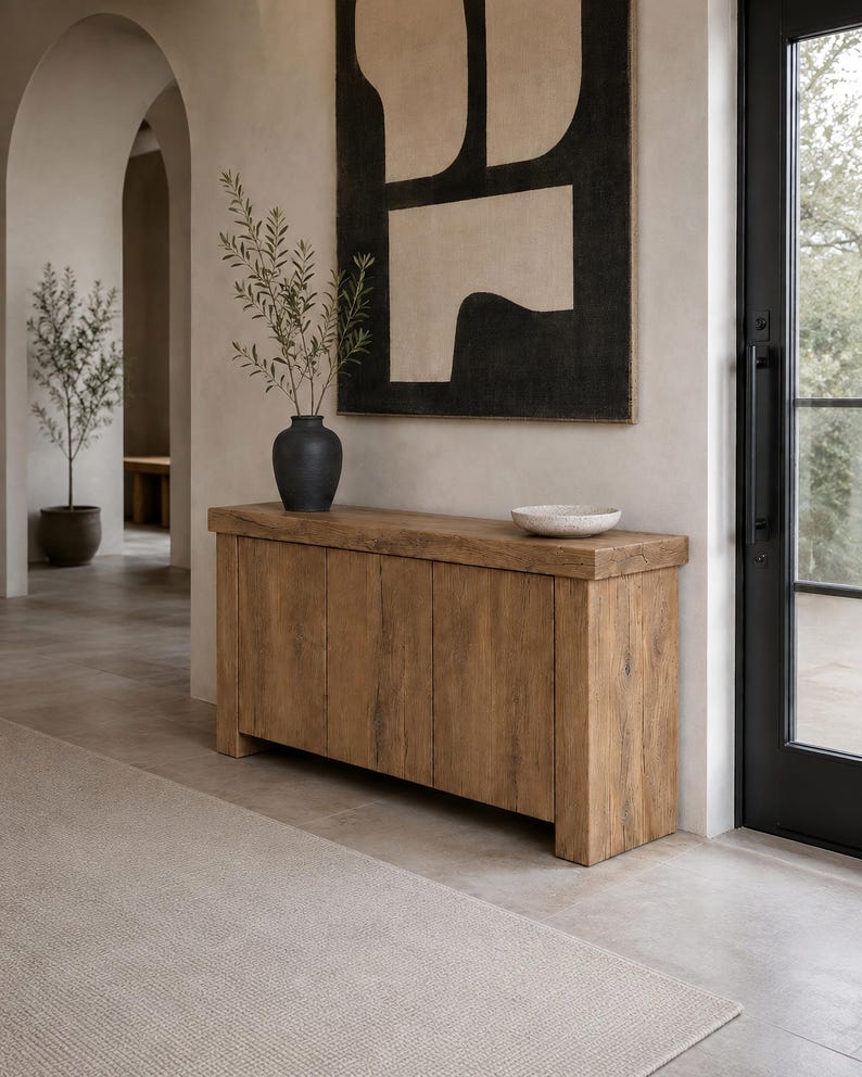 Peut inclure: Une console rectangulaire en bois avec une finition &agrave; grain naturel et quatre portes de placard. Sur le dessus se trouvent un vase noir mat contenant des branches feuillues vertes et un bol peu profond blanc cass&eacute; mouchet&eacute;.