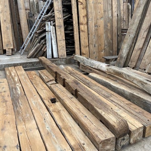 May include: A pile of reclaimed wood beams, stacked and ready for use in a construction project. The beams are weathered and have a rustic appearance.