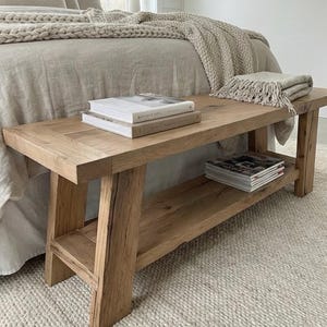 May include: A light-colored wooden bench with a lower shelf, holding books and magazines. The bench is placed at the foot of a bed with a neutral-toned comforter and a textured rug.