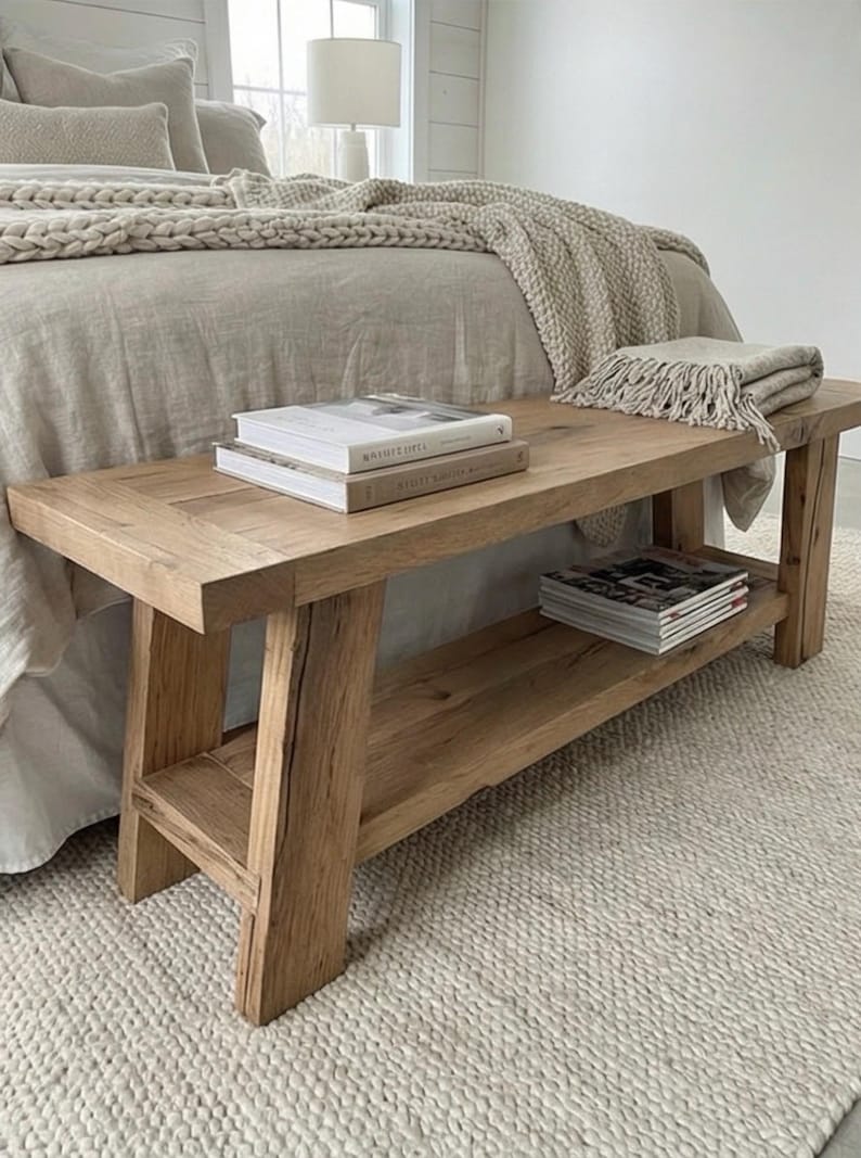 May include: A light-colored wooden bench with a lower shelf, holding books and magazines. The bench is placed at the foot of a bed with a neutral-toned comforter and a textured rug.