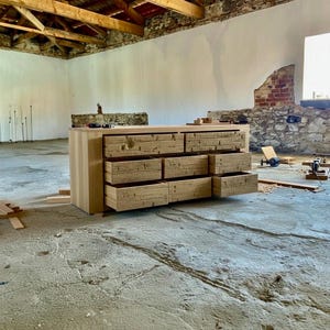 May include: A light-colored wooden dresser with eight drawers, some of which are open. The dresser is in a room with a concrete floor and exposed brick and stone walls. The wood has a natural grain.