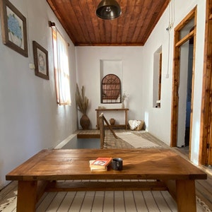 May include: A living room with a large, rectangular wooden coffee table. The room features a wooden ceiling, white walls, and a patterned rug. A book and a black mug sit on the table. Decorative elements include framed artwork and a hanging light fixture.