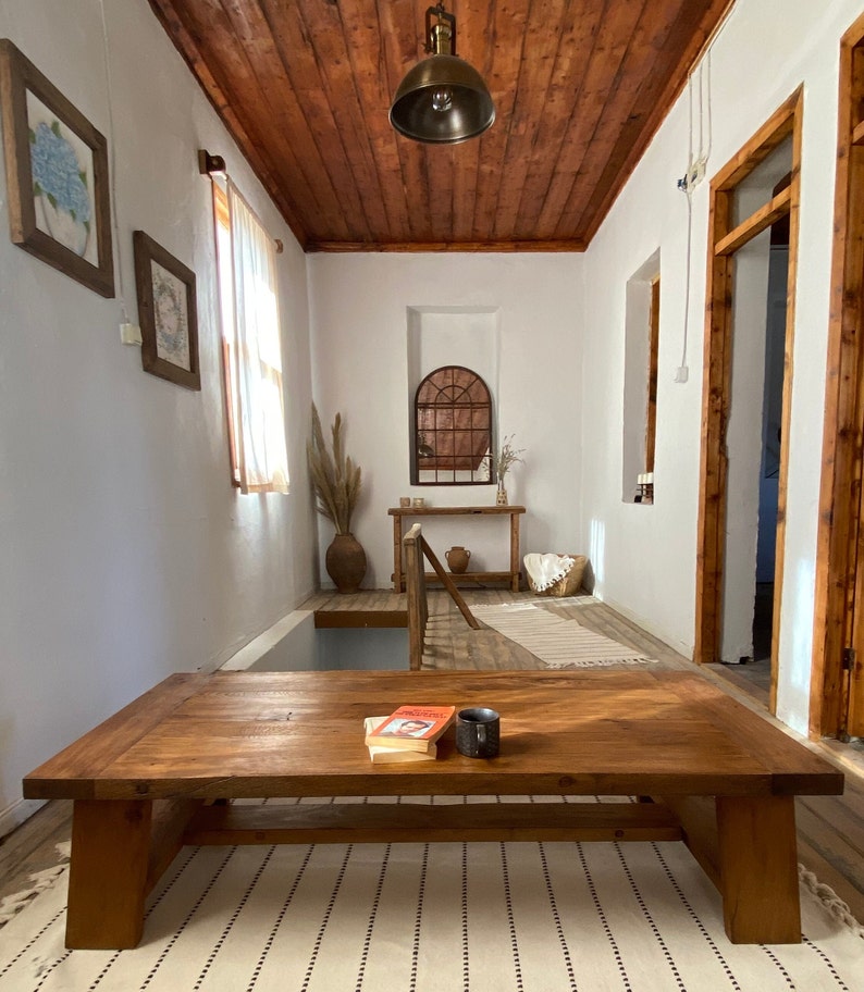 May include: A living room with a large, rectangular wooden coffee table. The room features a wooden ceiling, white walls, and a patterned rug. A book and a black mug sit on the table. Decorative elements include framed artwork and a hanging light fixture.