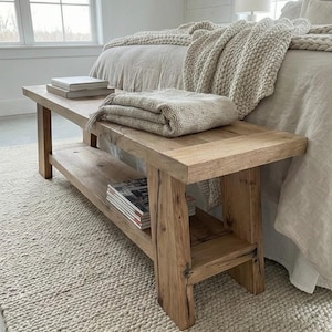 May include: A rustic wooden bench with a lower shelf, holding magazines. A folded beige blanket and two books rest on the bench. The bench sits on a cream-colored rug, near a bed with a beige blanket.