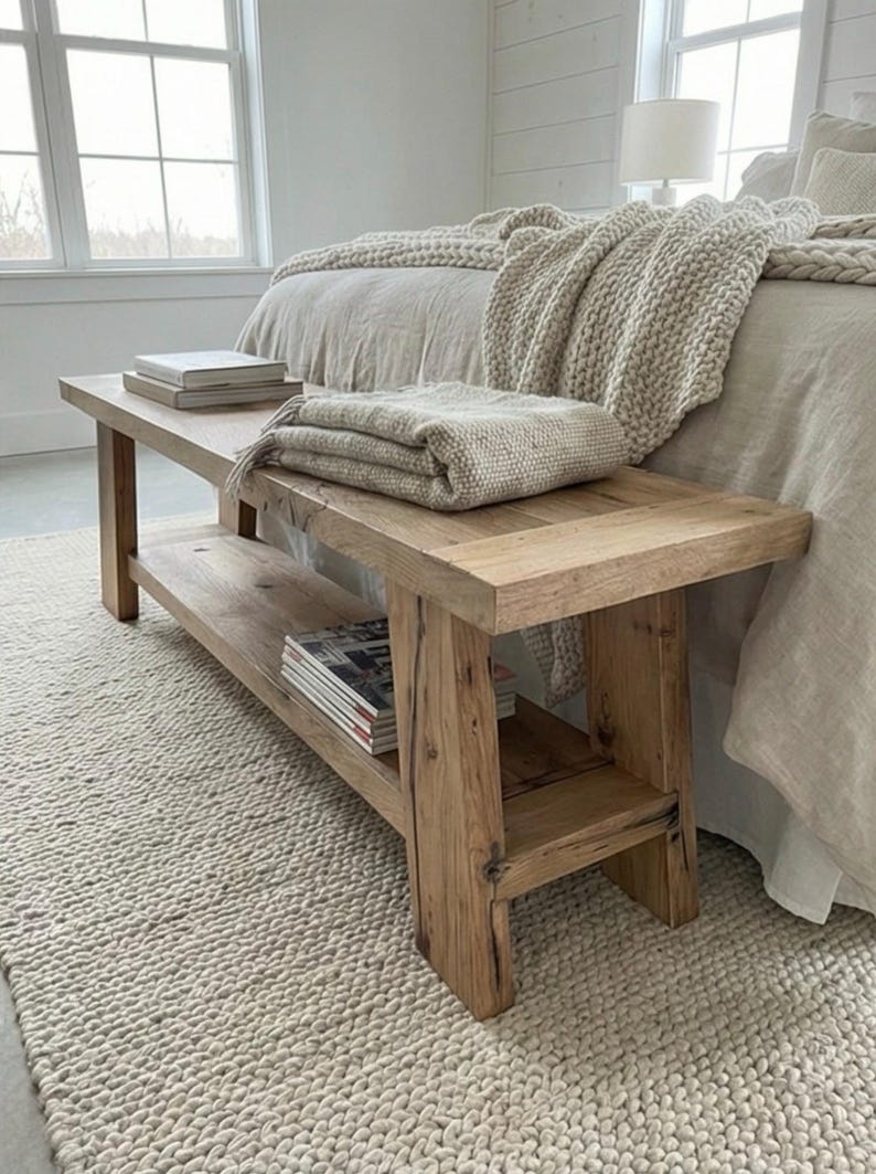 May include: A rustic wooden bench with a lower shelf, holding magazines. A folded beige blanket and two books rest on the bench. The bench sits on a cream-colored rug, near a bed with a beige blanket.