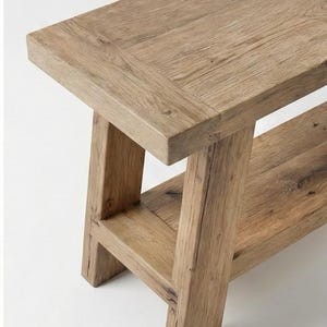 May include: A rustic wooden bench with a natural finish. The bench features a rectangular top and a lower shelf, supported by sturdy, angled legs. The wood grain is visible throughout, adding to its natural aesthetic.