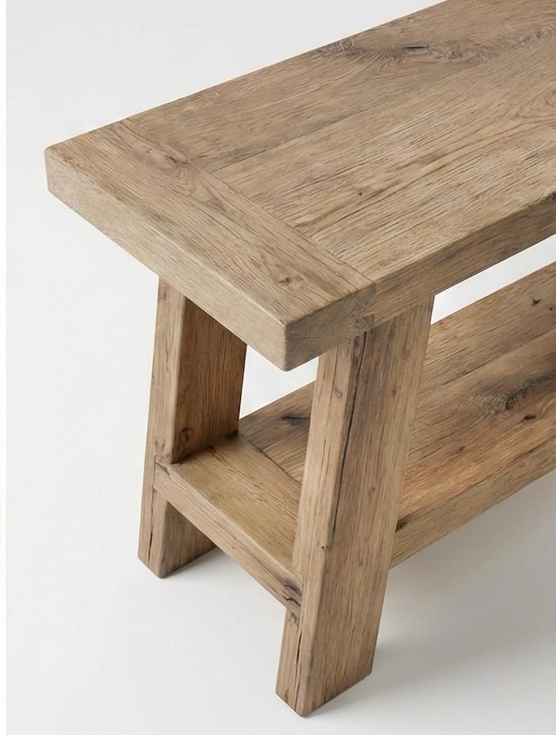 May include: A rustic wooden bench with a natural finish. The bench features a rectangular top and a lower shelf, supported by sturdy, angled legs. The wood grain is visible throughout, adding to its natural aesthetic.