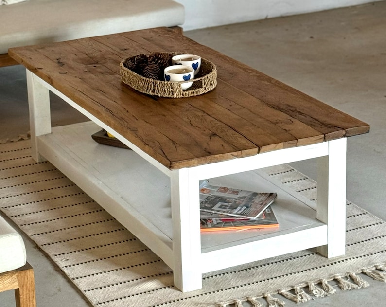 May include: A white coffee table with a wooden top and a lower shelf. The table has a rustic, farmhouse style and is decorated with a woven basket and three small cups.