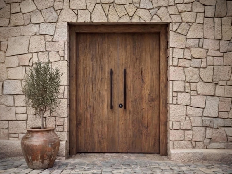 May include: A set of double wooden doors with a natural brown finish and vertical grain, featuring two long, thin, black metal vertical handles and a central keyhole, set within a wall constructed of light-colored, irregular-shaped stone blocks.