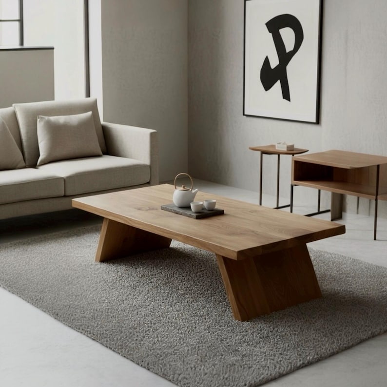 Puede incluir: Un sal&oacute;n moderno con una gran mesa de centro rectangular de madera con patas anguladas. Una tetera y tazas blancas est&aacute;n sobre una bandeja oscura. Un sof&aacute; y una alfombra de color gris claro complementan la mesa. Una obra de arte abstracta enmarcada cuelga en la pared.