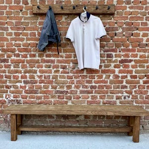 May include: A wooden bench with a shelf underneath. The bench is made of dark brown wood and has a rustic look. The bench is in front of a brick wall with a wooden coat rack above it. A white polo shirt and a pair of blue jeans are hanging on the coat rack.