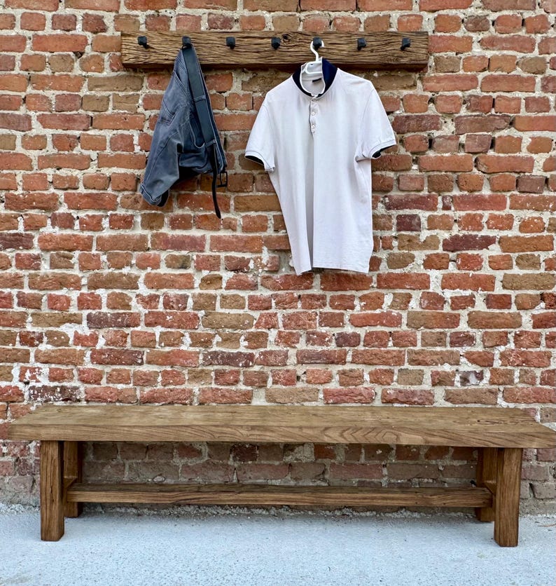May include: A wooden bench with a shelf underneath. The bench is made of dark brown wood and has a rustic look. The bench is in front of a brick wall with a wooden coat rack above it. A white polo shirt and a pair of blue jeans are hanging on the coat rack.