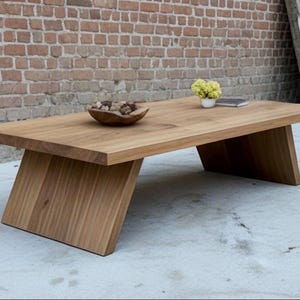 Puede incluir: Una mesa de centro rectangular de madera con patas anguladas. La mesa es de madera clara y tiene un acabado natural. Un cuenco de madera con piedras decorativas y un peque&ntilde;o jarr&oacute;n con flores amarillas est&aacute;n sobre la mesa.