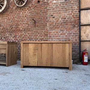 Peut inclure: Un buffet rectangulaire en bois dot&eacute; de trois portes verticales avec un grain de bois naturel visible. Ce meuble pr&eacute;sente une surface sup&eacute;rieure plane et une structure robuste, pos&eacute; sur un sol en b&eacute;ton devant un mur en briques.