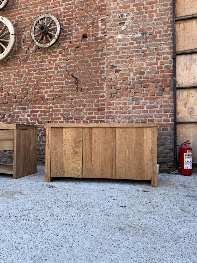 Peut inclure: Un buffet rectangulaire en bois dot&eacute; de trois portes verticales avec un grain de bois naturel visible. Ce meuble pr&eacute;sente une surface sup&eacute;rieure plane et une structure robuste, pos&eacute; sur un sol en b&eacute;ton devant un mur en briques.