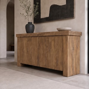 Peut inclure: Un buffet rectangulaire en bois avec une finition brun clair naturelle, mettant en valeur le grain et les n&oelig;uds du bois. Le meuble poss&egrave;de quatre portes verticales, un plateau &eacute;pais et des pieds blocs robustes. Un vase sombre et un bol sont pos&eacute;s dessus.