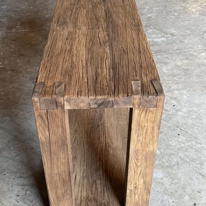 K&ouml;nnte beinhalten: Ein rustikaler Holz-Konsolentisch mit einer nat&uuml;rlichen braunen Oberfl&auml;che. Der Tisch hat ein einzelnes Regal und ist aus recyceltem Holz gefertigt.