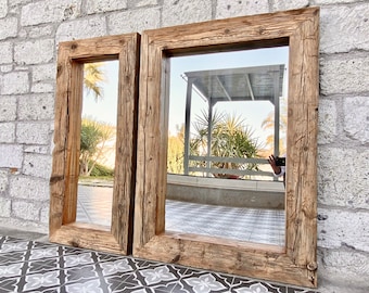 May include: Two rectangular mirrors with light brown, rustic wooden frames. The frames have a rough, textured appearance. The mirrors reflect an outdoor scene with a gazebo and greenery. The mirrors are leaning against a light-coloured brick wall.