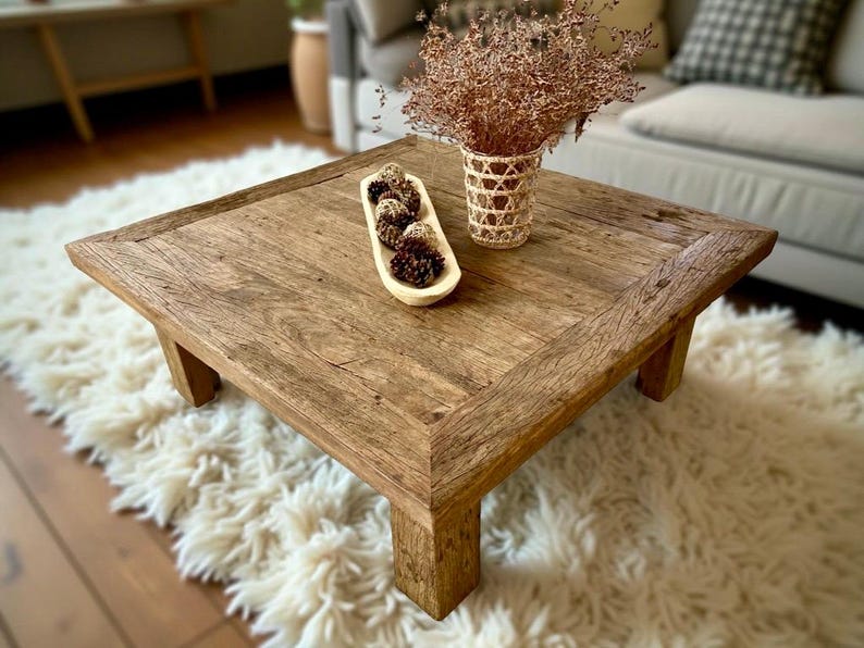 May include: Square wooden coffee table with a natural finish. The table has a rustic design with visible wood grain and sturdy legs. A woven vase with dried flowers and a wooden bowl with pinecones are on the table. The table sits on a fluffy white rug.