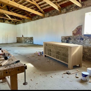 May include: A light-colored wooden dresser with multiple drawers and a natural wood finish. The dresser is in a workshop setting, with tools and paint cans nearby. The room has exposed beams and stone walls.