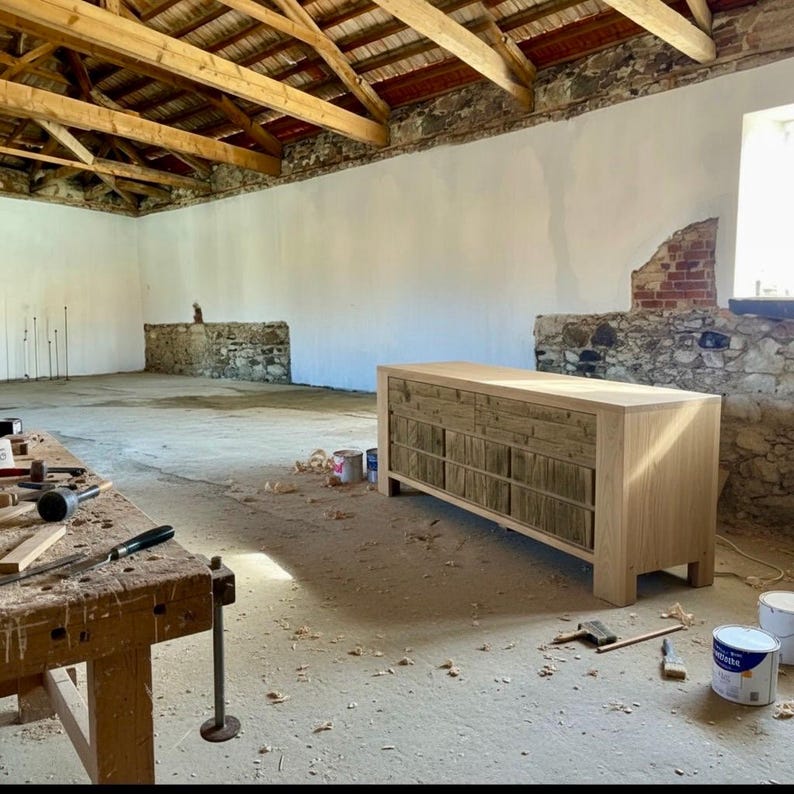 May include: A light-colored wooden dresser with multiple drawers and a natural wood finish. The dresser is in a workshop setting, with tools and paint cans nearby. The room has exposed beams and stone walls.