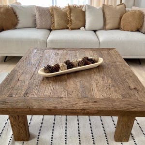 May include: A rustic, square wooden coffee table with a distressed finish. A wooden bowl filled with decorative pine cones and spheres sits on the table. A cream-colored sofa with decorative pillows is in the background.