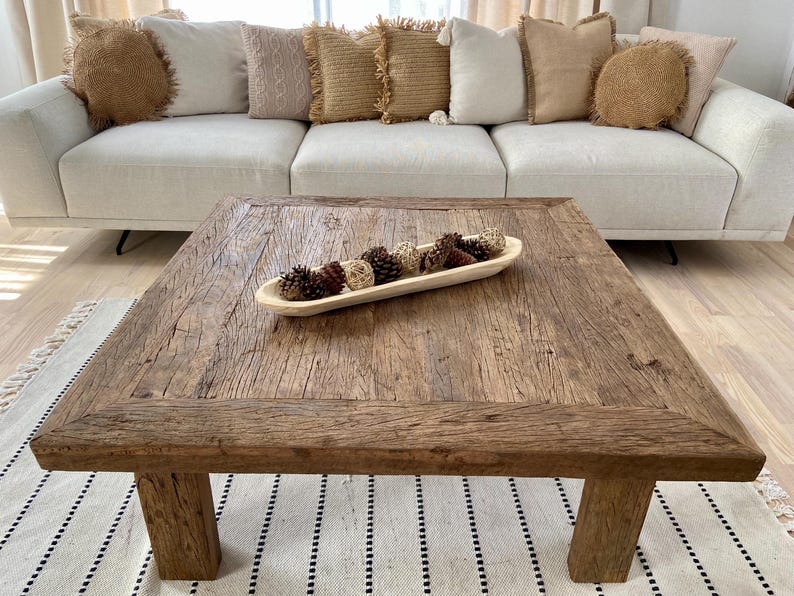 May include: A rustic, square wooden coffee table with a distressed finish. A wooden bowl filled with decorative pine cones and spheres sits on the table. A cream-colored sofa with decorative pillows is in the background.