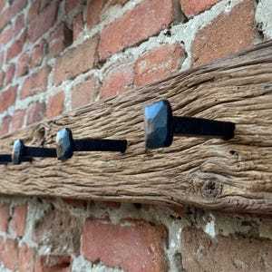 May include: A rustic wooden coat rack with six black metal hooks. The rack is mounted on a brick wall.
