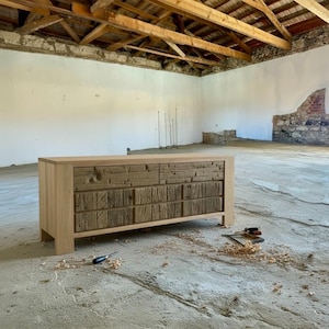 May include: A light-colored wooden dresser with two rows of drawers. The top row has two drawers, and the bottom row has four drawers. The drawer fronts have a textured, rustic appearance. The dresser is in a large, open room with a concrete floor.