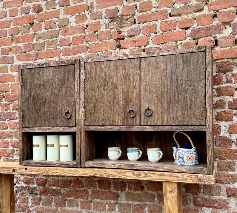 Puede incluir: Un armario de madera con dos puertas y un estante. El armario tiene dos peque&ntilde;os recipientes blancos en el estante y tres tazas peque&ntilde;as y una tetera con un patr&oacute;n floral en el estante inferior. El armario est&aacute; montado en un estante de madera contra una pared de ladrillo.