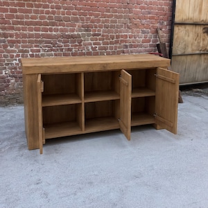 Peut inclure: Un buffet rectangulaire en bois avec une finition naturelle en grain de bois brun clair. Le meuble poss&egrave;de un plateau &eacute;pais et trois sections de rangement ouvertes, chacune divis&eacute;e en deux niveaux par une &eacute;tag&egrave;re, avec deux portes battantes.