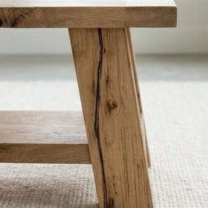 May include: A light-colored wooden bench with a rectangular top and angled legs. The bench has a lower shelf and is made of natural wood with visible grain and a dark crack detail on one leg.