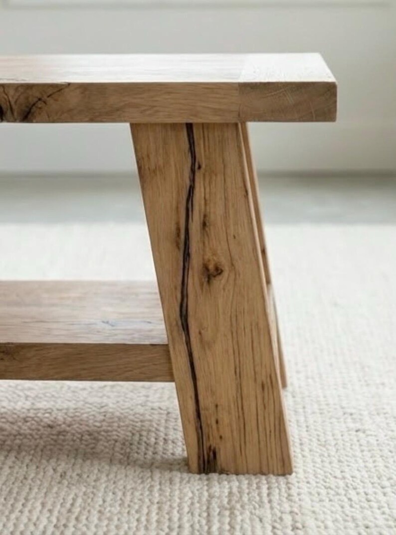 May include: A light-colored wooden bench with a rectangular top and angled legs. The bench has a lower shelf and is made of natural wood with visible grain and a dark crack detail on one leg.
